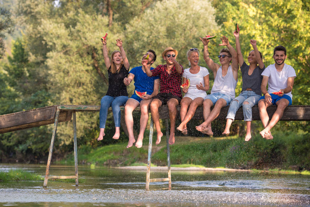 Grupa młodych przyjaciół ciesząc się arbuz siedząc na drewniany most nad rzeką w pięknej przyrody - Zdjęcie, obraz