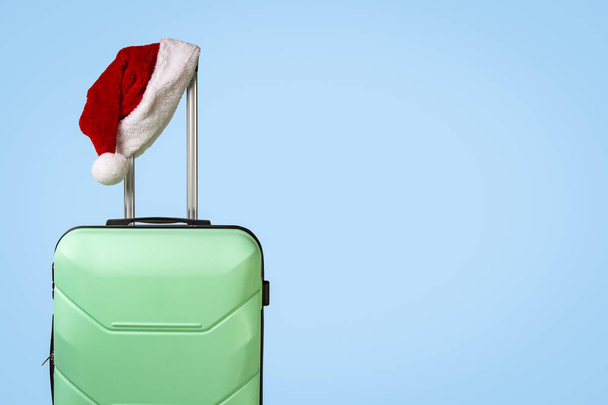 Plastic suitcase and Santa Claus hat on blue background. Concept of travel to visit friends and relatives on Christmas holidays. Merry Christmas and Happy New Year. Christmas trip. - Photo, image