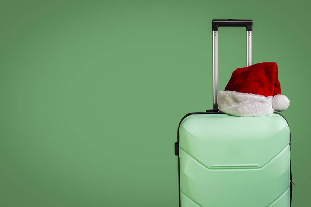 Plastic suitcase and Santa Claus hat on green background. Concept of travel to visit friends and relatives on Christmas holidays. Merry Christmas and Happy New Year. Christmas trip. - Photo, image
