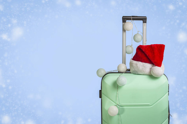 Plastic suitcase, Santa Claus cap and garland on a blue background with snow. Concept of travel, business trips, trips to visit friends and relatives on Christmas holidays. New Year's journey. - Fotografie, Obrázek
