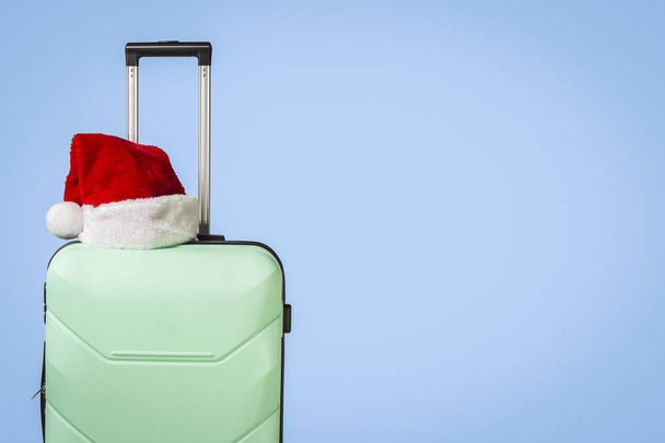 Plastic suitcase and Santa Claus hat on blue background. Concept of travel to visit friends and relatives on Christmas holidays. Merry Christmas and Happy New Year. Christmas trip. - Foto, imagen
