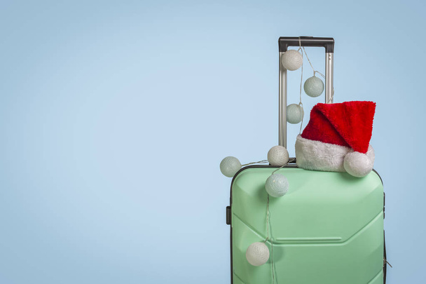 Plastic suitcase, Santa Claus cap and garland on a blue background. Concept of travel, business trips, trips to visit friends and relatives on Christmas holidays. New Year's journey. - Фото, зображення
