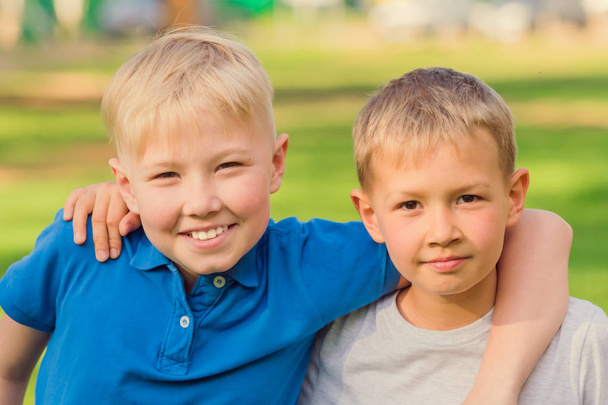 Two blond boys in white and blue T-shirts standing embracing in summer park - Photo, image