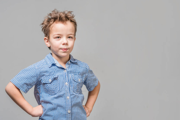 Cute little boy in a blue shirt standing with his hands on his hips on a gray background. Isolated - Zdjęcie, obraz