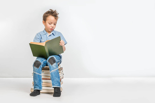 Cute boy in a blue shirt with a book sitting on a pile of books - Фото, изображение