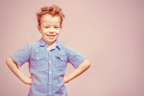 Cute little boy in a blue shirt standing with his hands on his hips on a pink background. Toned - Foto, Imagem