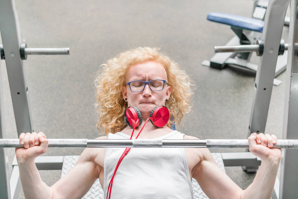 Curly blond man in a white vest lifting barbell with a strained face in the gym - Foto, Imagem