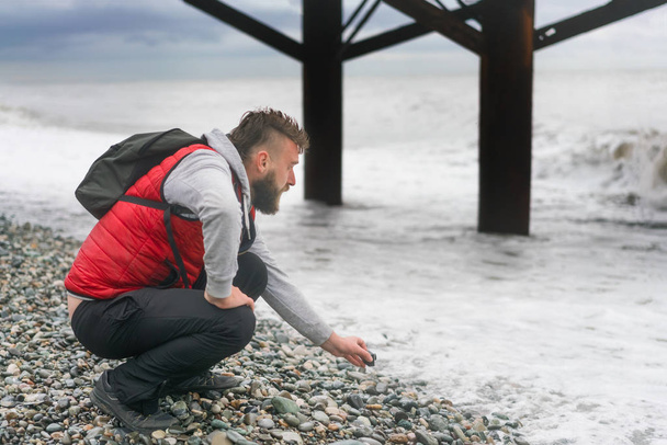 Man in a red vest and backpack sitting near the sea on a pebble beach - Foto, Bild