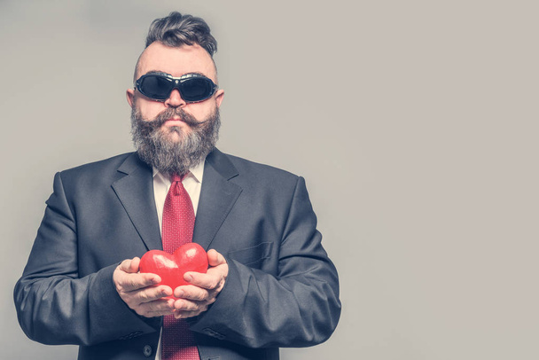 Adult bearded man in a jacket, red tie and sunglasses holding a red heart on a gray background - Foto, Imagen