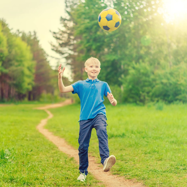 Blond boy in a blue T-shirt kicking a ball on a forest path in the sunlight - Fotó, kép