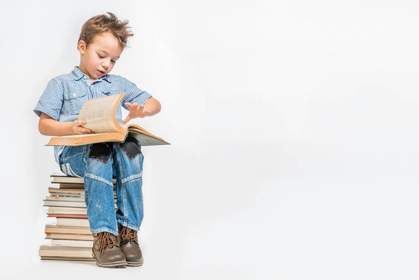 Roztomilý chlapec s knihou sedí na hromadě knih na bílém pozadí. Izolovaný - Fotografie, Obrázek