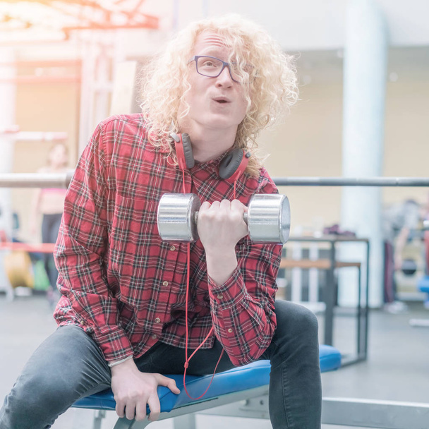 Curly blond man with glasses raises a small dumbbell in the gym with a strained face - Photo, Image