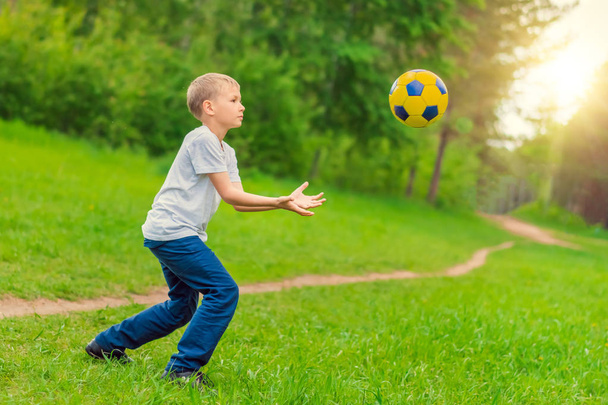 Blond boy in a white T-shirt playing with a ball near the path in the park - Φωτογραφία, εικόνα