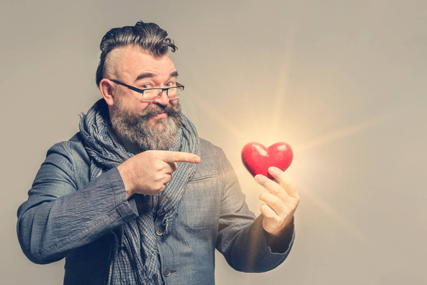 Adult bearded man in a blue jacket and scarf pointing at red heart - Fotografie, Obrázek