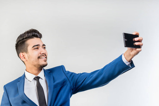 Young attractive man in a blue suit looking at the phone's screen on a gray background. Isolated - Foto, imagen