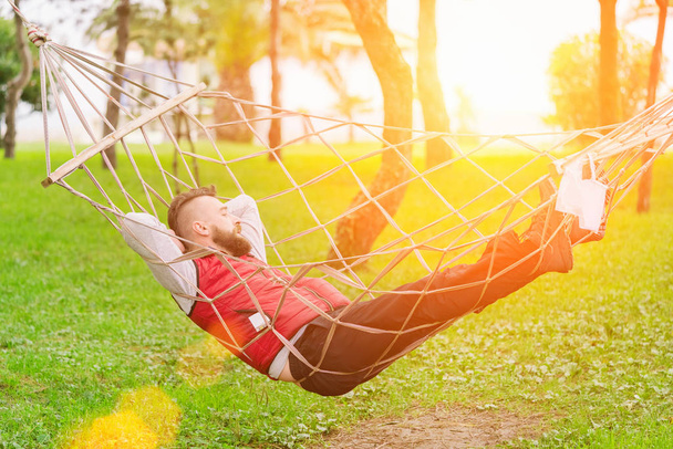Parrakas mies punaliivissä nukkuu riippumatossa puistossa aurinkoisena päivänä.
 - Valokuva, kuva
