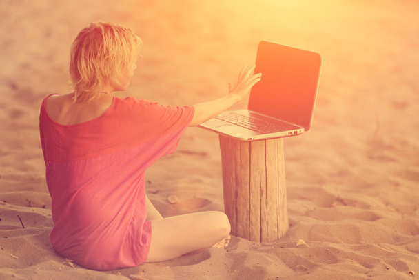 Girl blonde teenager in red blouse sitting in front of a laptop on the beach at sunset - 写真・画像