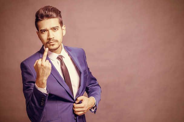 Young attractive man in a blue suit showing middle finger on brown background - Photo, Image