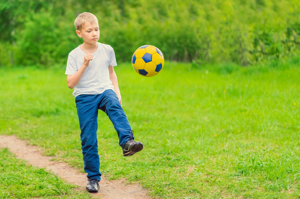 Blond boy in a white T-shirt walking along the path and kicking a yellow soccer ball. Toned - 写真・画像