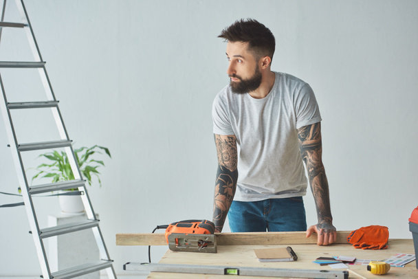 handsome bearded man leaning at wooden table and looking away during house repair  - Zdjęcie, obraz
