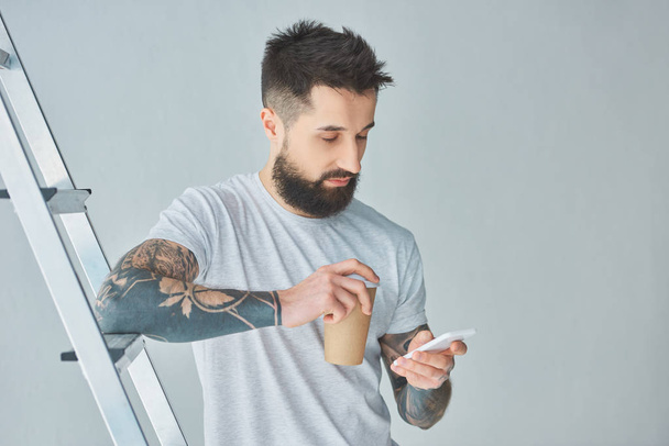 bearded young man holding paper cup and using smartphone while standing near stepladder on grey - Foto, imagen