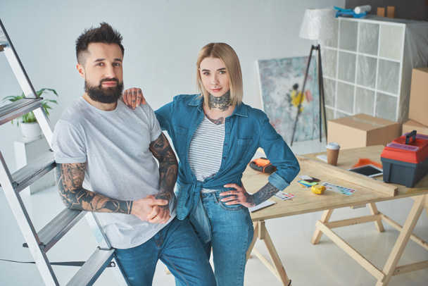 kaunis nuori tatuoitu pari katselee kameraa seisoessaan yhdessä uudessa kodissa
 - Valokuva, kuva