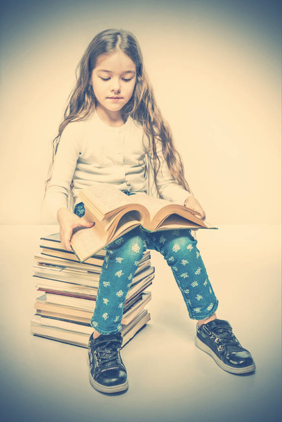 Cute girl with a book sitting on a pile of books. Toned - Φωτογραφία, εικόνα