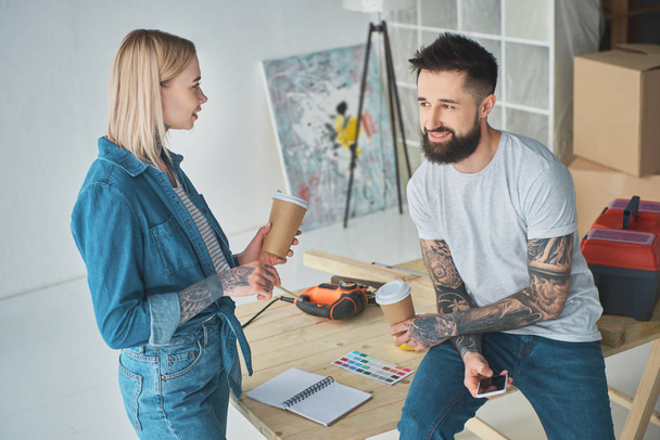 happy young couple drinking coffee from paper cups and using smartphone during home improvement - Photo, Image