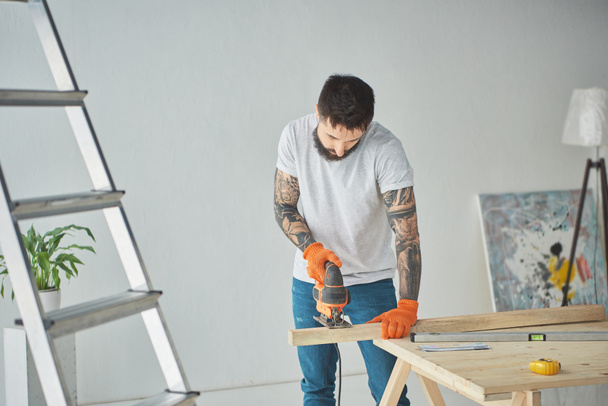 handsome bearded tattooed man using electric jigsaw during house repair - Photo, Image