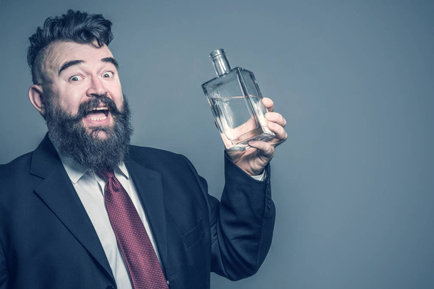 Crazy bearded man in suit holding a bottle of alcohol on a gray background. Toned - Фото, изображение