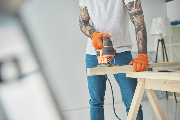 cropped shot of young tattooed man using electric jigsaw during home improvement - Foto, Bild