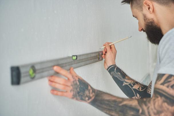 cropped shot of handsome bearded young man holding level tool and pencil during home improvement - Fotoğraf, Görsel