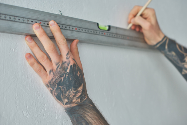 περικοπεί shot του τατουάζ άνθρωπος που κρατά επίπεδο εργαλείο και σήμανσης τοίχο με μολύβι κατά τη διάρκεια επισκευών - Φωτογραφία, εικόνα