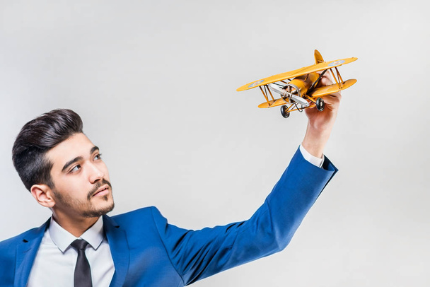 Young attractive man in a blue suit holding a yellow airplane model on a gray background. Isolated - Photo, Image