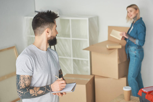bearded man taking notes and looking at girlfriend unpacking cardboard boxes in new house - Foto, Imagem