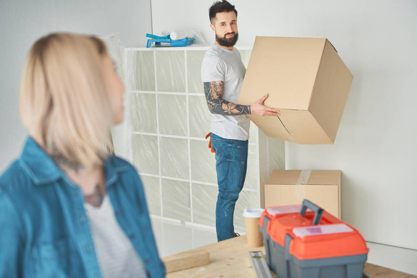 mujer joven mirando novio barbudo sosteniendo caja de cartón en casa nueva
 - Foto, Imagen