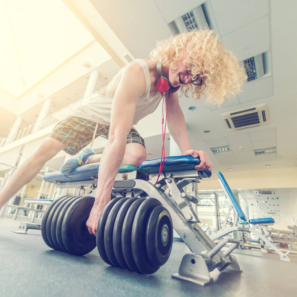 Weak curly blonde man trying to raise heavy dumbbell in the gym in the sunlight - Photo, Image