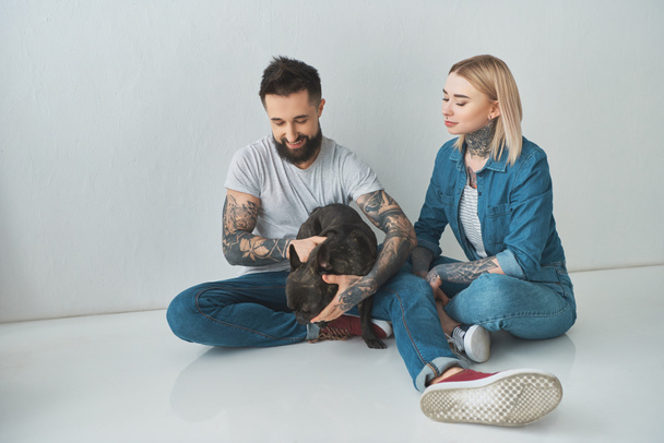 happy young couple sitting and playing with french bulldog on grey - Zdjęcie, obraz