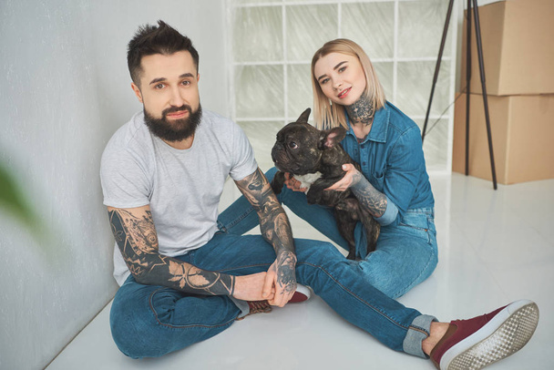 onnellinen nuori tatuoitu pari leikkii ranskalaisen bulldogin kanssa ja hymyilee kameralle uudessa kodissa
 - Valokuva, kuva