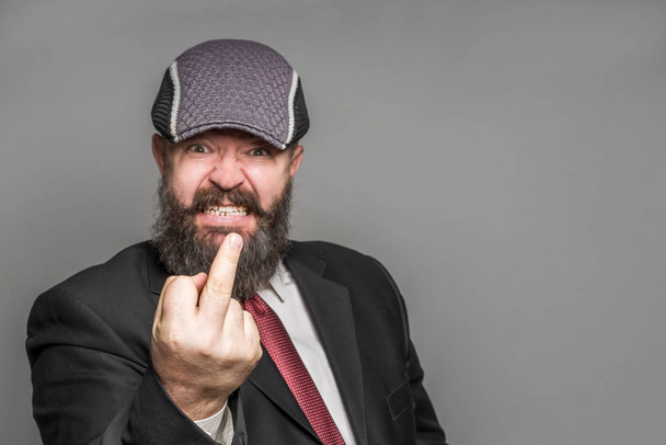 Angry adult bearded man in a suit and cap showing middle finger on a gray background - Foto, imagen