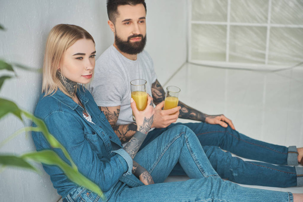 pareja joven sosteniendo vasos de jugo y mirando hacia otro lado mientras se sienta en el piso en una casa nueva
 - Foto, imagen