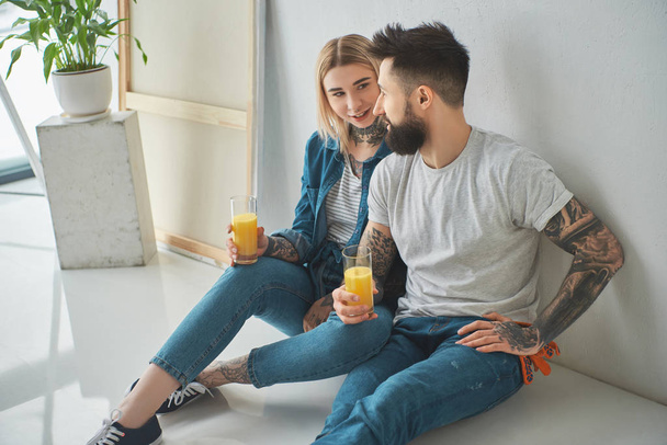 happy young couple holding glasses of juice and sitting on floor in new home - Foto, afbeelding