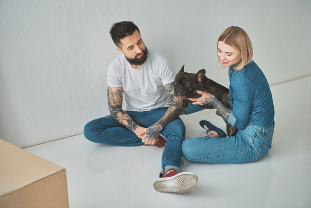 Blick aus der Vogelperspektive auf lächelnde junge tätowierte Paare, die auf dem Boden sitzen und im neuen Haus mit Hund spielen - Foto, Bild