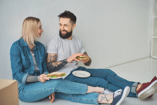 happy young couple holding sandwiches and smiling each other in new house  - Photo, Image