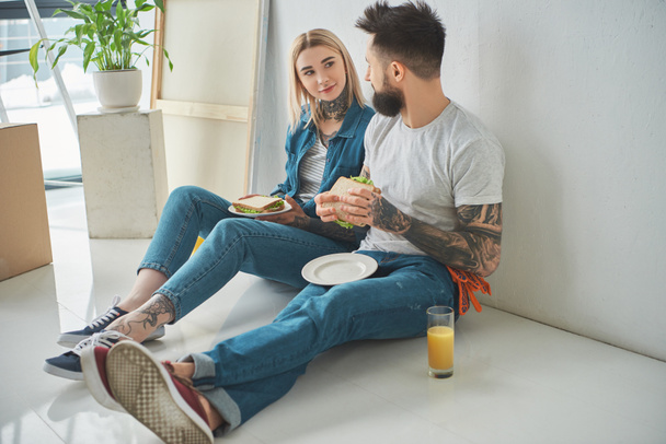 young tattooed couple eating sandwiches and looking at each other while sitting on floor i new house - Foto, afbeelding