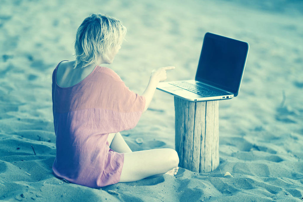 Dívka blondýna teenager v červená halenka sedí v přední části notebooku na pláži. Tónovaný - Fotografie, Obrázek