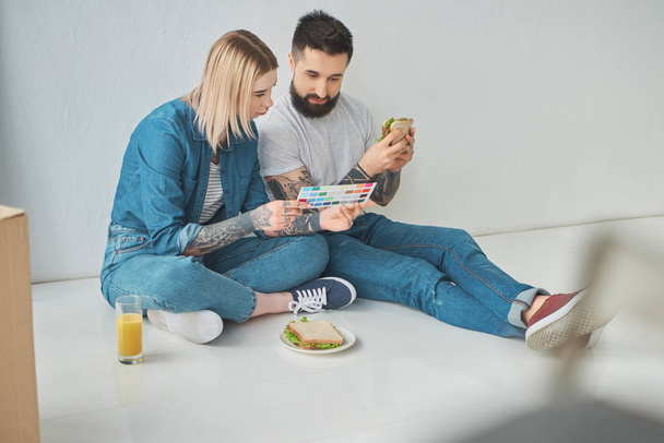 vista de ángulo alto de la pareja comiendo sándwiches y mirando la paleta de colores en un nuevo hogar
 - Foto, imagen