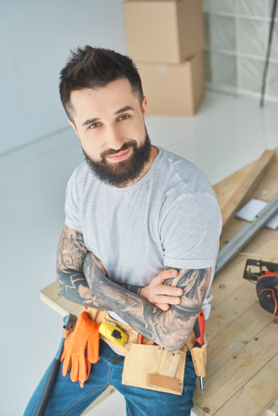 smiling repairman with tattoos and tools leaning on wooden surface in new apartment - Fotografie, Obrázek