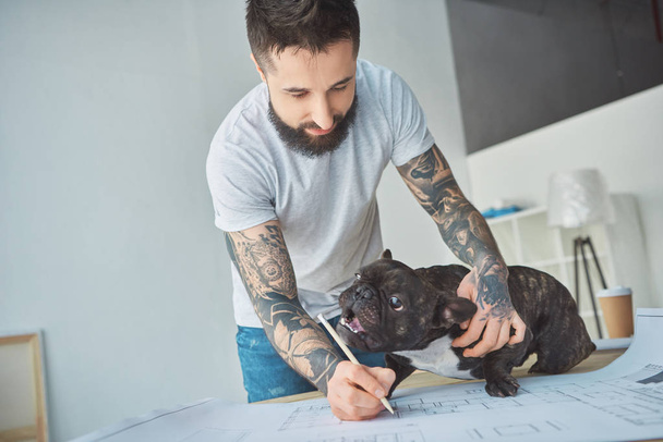 reparador tatuado con lápiz y bulldog francés mirando el plano de nuevo apartamento en la mesa de madera
 - Foto, Imagen