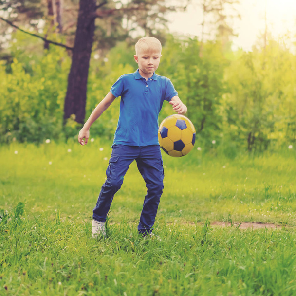 Blond boy in a blue T-shirt kicking the ball at the edge of the forest - Foto, Imagen